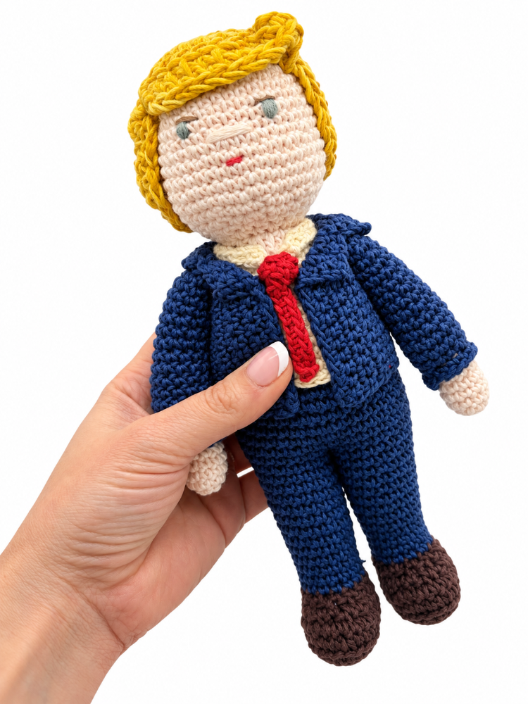 Handmade crochet Trump doll held in one hand, showing yellow hair, a navy suit, red tie and brown shoes.