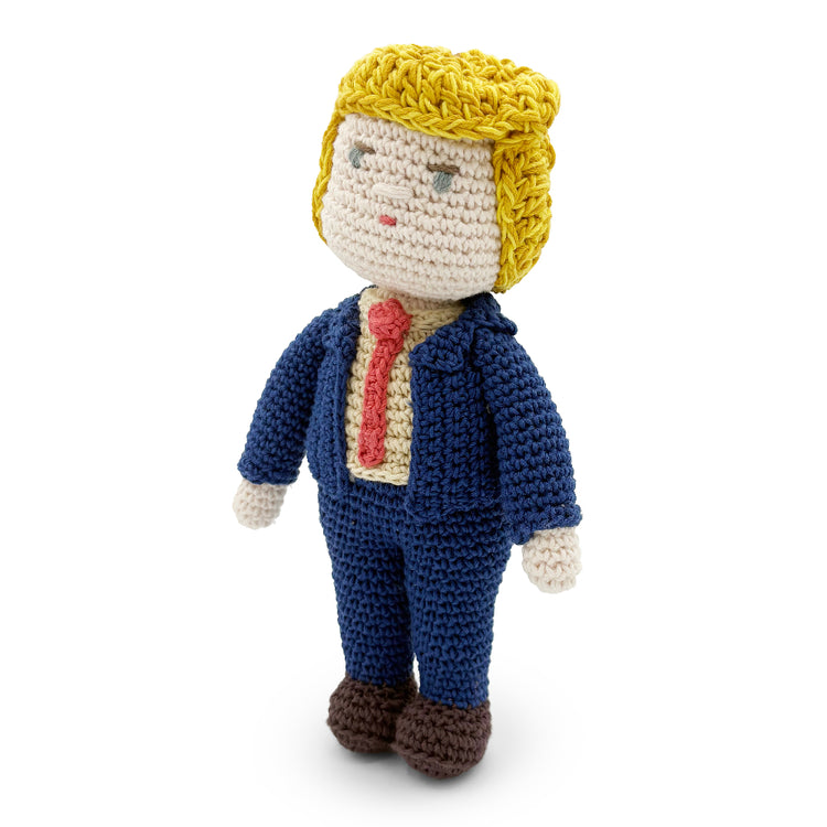 Three-quarter view of a crochet Trump doll showing yellow yarn hair, a navy jacket, red tie and brown shoes.